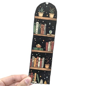 May include: A black bookmark featuring a bookshelf design. The bookshelf is filled with books, plants, and crystals. The bookmark is shaped like a tall, narrow rectangle.