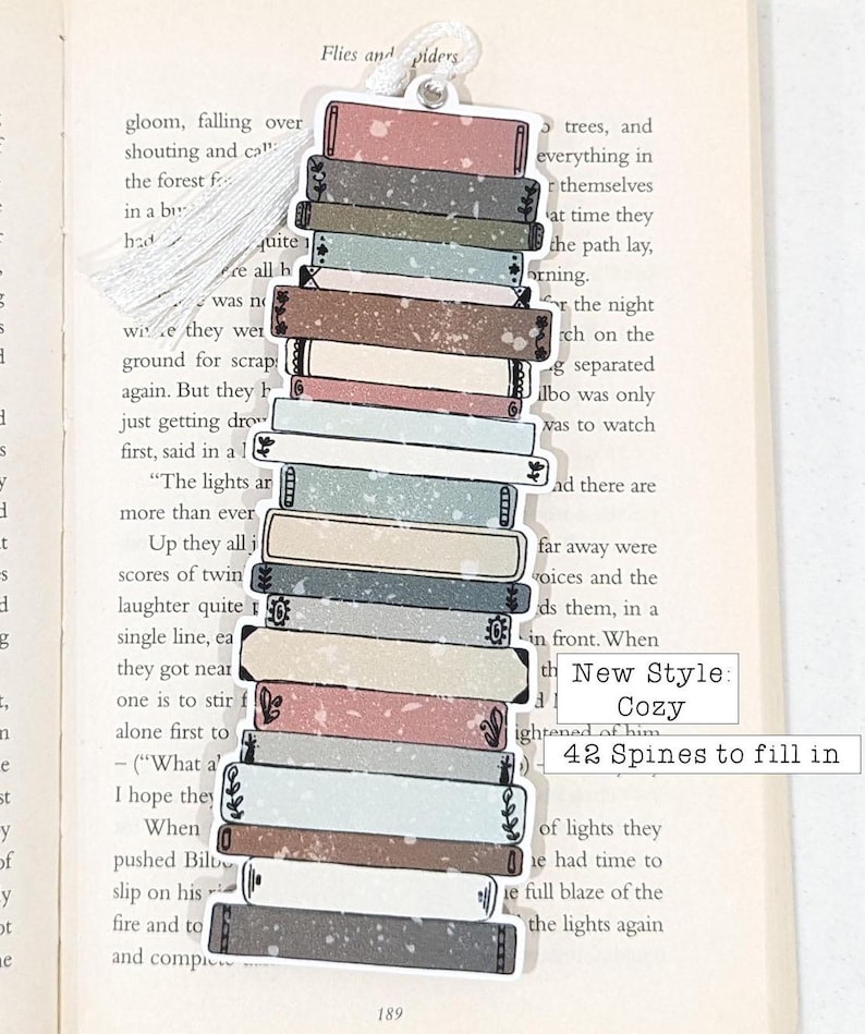 Puede incluir: Una pila de 42 lomos de libros con un marcap&aacute;ginas de borla blanca. El marcap&aacute;ginas est&aacute; decorado con una pila de libros de varios colores y patrones. El texto "New Style: Cozy" y "42 Spines to fill in" est&aacute; impreso en el marcap&aacute;ginas.