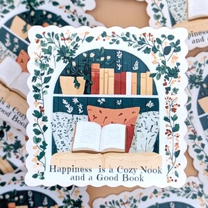 Puede incluir: Una pegatina que representa una escena de rinc&oacute;n de lectura. La ilustraci&oacute;n muestra una ventana con una estanter&iacute;a, un libro abierto y cojines. El follaje verde enmarca la escena, y el texto "Happiness is a Cozy Nook and a Good Book" est&aacute; en la parte inferior.