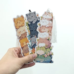 May include: Two colourful bookmarks featuring stacks of cute cartoon cats. The bookmarks have tassels and are perfect for book lovers.