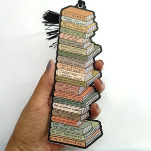 May include: A bookmark featuring a stack of books with the titles of popular novels. The bookmark is made of sturdy cardboard and has a tassel.