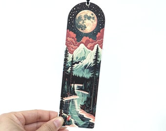 May include: A bookmark featuring a landscape scene with a full moon, mountains, and a river. The design includes a dark night sky with stars, red clouds, and evergreen trees. The bookmark has a hole at the top for a string.