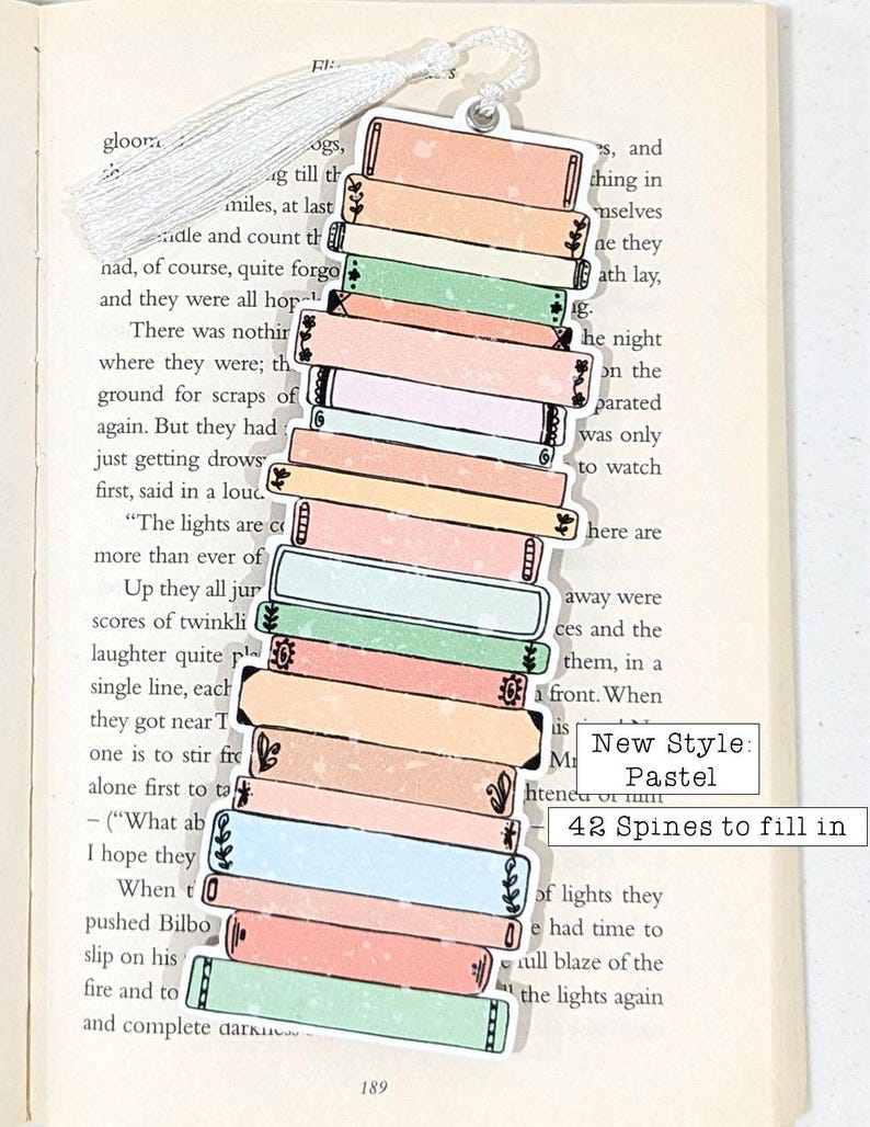 Puede incluir: Un marcap&aacute;ginas con forma de pila de libros con lomos de colores pastel. El marcap&aacute;ginas tiene un borla blanca y el texto "New Style: Pastel 42 Spines to fill in" est&aacute; impreso en la parte inferior.