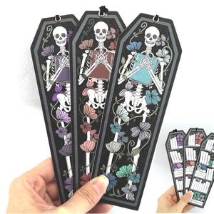 May include: Three black coffin-shaped bookmarks featuring a skeleton holding a book and surrounded by colourful flowers. The skeletons are holding different coloured books: purple, brown, and blue.