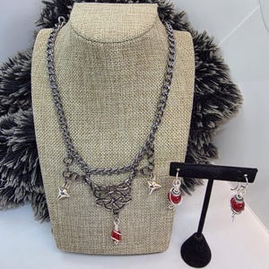 May include: A gunmetal-toned necklace and earring set. The necklace features a chain design with Celtic knotwork and red gemstone accents. The earrings have matching red gemstone drops. The jewelry is displayed on a mannequin.