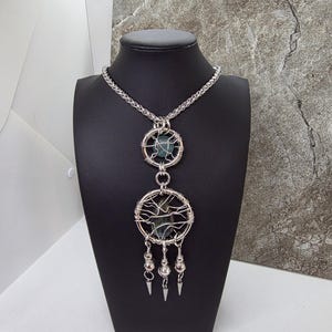 May include: A silver-toned wire-wrapped pendant necklace. The necklace features two circular pendants, each with a central stone. The lower pendant has dangling silver accents. The necklace is displayed on a black bust.