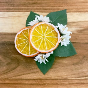 May include: Two orange slices with a white rim, arranged on a bed of green leaves and white flowers.