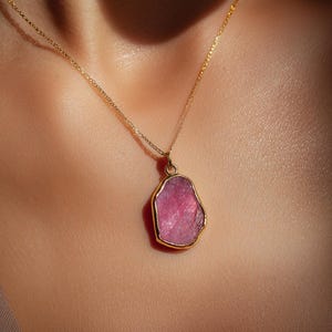 May include: A gold necklace with a pink gemstone pendant. The pendant has a unique, irregular shape and is set in a gold bezel. The necklace is worn against a neutral-toned background, highlighting the jewelry's color and design.