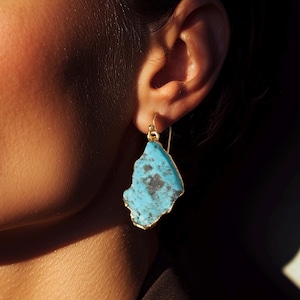 May include: A gold earring featuring a turquoise stone with a rough, irregular shape. The stone is set within a gold border and hangs from a gold hook. The image is a close-up, with a person's face and shoulder in the blurred background.