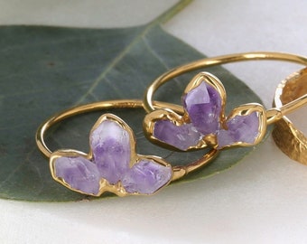 May include: Two gold rings featuring clusters of raw, purple amethyst crystals and one plain gold band with a textured, hammered surface. The amethyst stones are set in gold metal, creating a floral-like shape on the thin, polished gold bands.