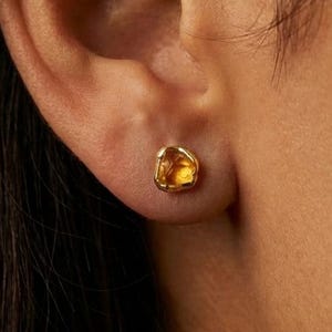 May include: A small, gold-toned stud earring with an irregular, organic shape. The center features a translucent, amber-colored stone set within a textured, metallic border that mimics the appearance of molten metal, secured to the earlobe.