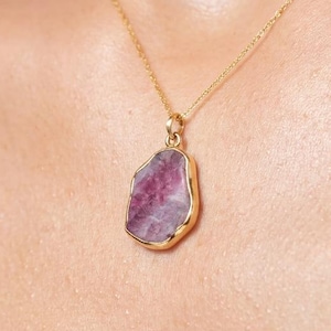May include: A gold chain necklace featuring a raw, irregular-shaped pinkish-purple gemstone pendant. The stone has a natural, unpolished texture and is encased in a thin gold-toned metal bezel setting that follows the organic contour of the gem.