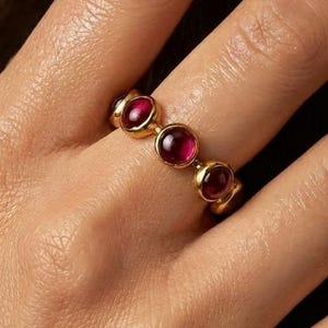 Puede incluir: Un anillo de oro con tres piedras preciosas redondas de color rojo intenso. El anillo tiene un diseño sencillo y elegante, que resalta el color vibrante de las piedras. El anillo se muestra en una mano, destacando su tamaño y estilo. Una joya.