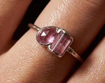Può includere: Un anello in argento con due pietre preziose rosa incastonate in una fascia di metallo. Una pietra è rotonda e sfaccettata, mentre l'altra è rettangolare e allungata. Le pietre sono posizionate fianco a fianco in una montatura d'argento sottile.