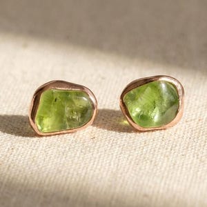 May include: A pair of stud earrings featuring raw, irregular-shaped green peridot gemstones set in polished rose gold metal settings. The stones have a translucent quality with natural inclusions, showcasing a vibrant lime green color.