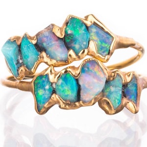 May include: A gold bypass ring featuring two rows of raw, uncut opals. The stones display iridescent flashes of blue, green, and pink. The metal band has a textured, organic finish that holds the irregular, jagged-edged opal gemstones in place.