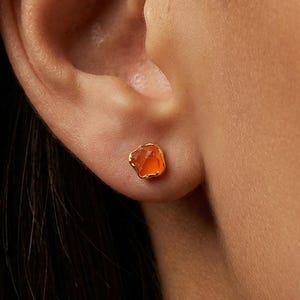 May include: Close-up of a single orange gemstone stud earring set in gold. The earring is a small, irregular shape, and is set against the skin of a person's ear.