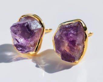 May include: A pair of stud earrings featuring raw, jagged-edged purple amethyst crystals. Each crystal is encased in a smooth, polished gold-toned metal setting that follows the irregular shape of the stone, highlighting the natural texture.