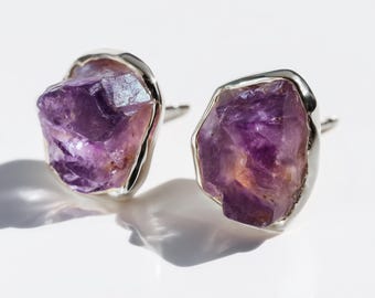 May include: A pair of stud earrings featuring raw, irregular amethyst crystals in shades of deep purple. Each stone is encased in a smooth, polished silver-toned metal setting that follows the natural, jagged contours of the gemstone surface.