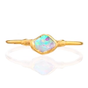 Puede incluir: Un anillo de oro con una piedra preciosa de ópalo en bruto, central e irregular, con destellos iridiscentes en azul, verde y rosa. La banda de metal tiene un acabado liso y pulido, y sostiene la piedra en un engaste que sigue su forma.