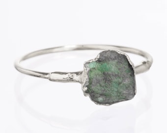 May include: A silver ring featuring a single, raw, uncut emerald gemstone. The stone has a rough, textured surface with deep green and dark grey inclusions. The metal band is thin and has a slightly irregular, organic finish where it meets the stone.