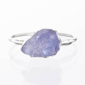 May include: A silver ring with a raw, light purple gemstone. The stone has an irregular shape and is set in a silver band with a textured design. The ring is presented on a white background, emphasising the natural beauty of the gemstone.