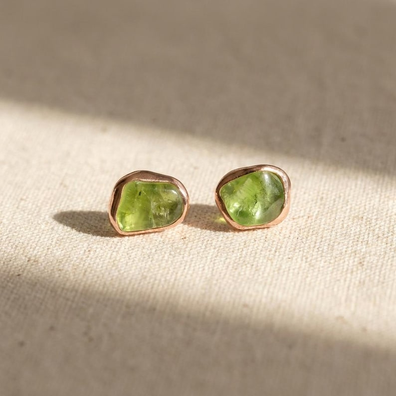 May include: A pair of stud earrings featuring raw, irregular-shaped green peridot gemstones set in polished rose gold metal settings. The stones have a translucent quality with natural inclusions, showcasing a vibrant lime green color.