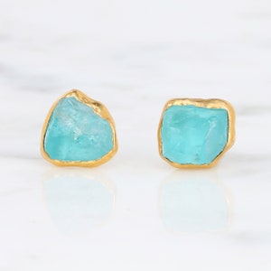 May include: A pair of stud earrings featuring raw, irregular turquoise-colored stones encased in a textured gold-toned metal setting. The stones have a translucent, crystalline appearance with jagged edges, contrasting with the smooth, polished gold border.