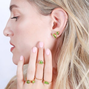 May include: A set of gold-toned jewelry featuring raw, light green peridot stones. The collection includes two stud earrings, a ring with a single stone, a ring with two stones, and a ring with a single stone, all set on thin gold bands.