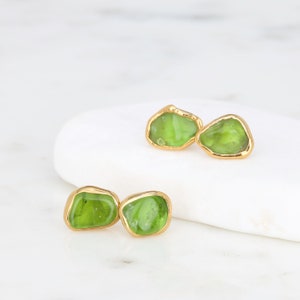 May include: A pair of stud earrings featuring irregular, translucent lime green stones encased in polished gold-toned metal settings. The stones have a smooth, organic shape with visible internal textures, and the gold frames follow the uneven contours of each individual stone.