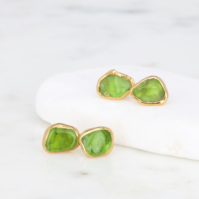 May include: A pair of stud earrings featuring irregular, translucent lime green stones encased in polished gold-toned metal settings. The stones have a smooth, organic shape with visible internal textures, and the gold frames follow the uneven contours of each individual stone.