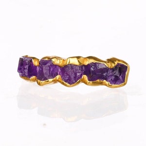May include: A gold ring featuring a row of five raw, deep purple amethyst stones set in a continuous metal band. The stones have an irregular, faceted texture, and the gold setting follows the natural contours of each individual gemstone.