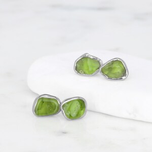 May include: A pair of stud earrings featuring irregular, translucent lime green stones encased in polished silver-toned metal settings. The stones have a smooth, organic shape with visible internal textures, contrasting with the smooth, metallic borders.