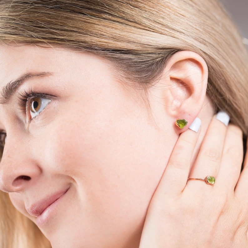 May include: A gold stud earring featuring a small, raw-cut, translucent lime green gemstone, paired with a matching thin gold ring set with a single, small, raw-cut, translucent lime green gemstone on a slender, polished gold band.