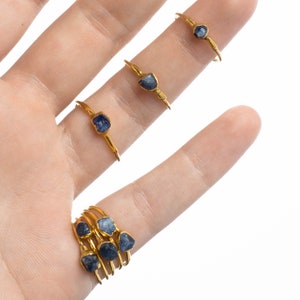 May include: A collection of gold-toned rings featuring raw, uncut blue gemstones. Three individual rings are worn on the upper fingers, while a stack of four rings with similar blue stones is worn on the base of the finger, all showcasing a metallic finish.