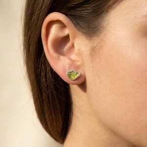 May include: A stud earring featuring a raw, irregular-shaped light green peridot gemstone set in a simple silver-toned metal bezel. The stone has a translucent, faceted appearance with natural inclusions and sits directly against the earlobe.