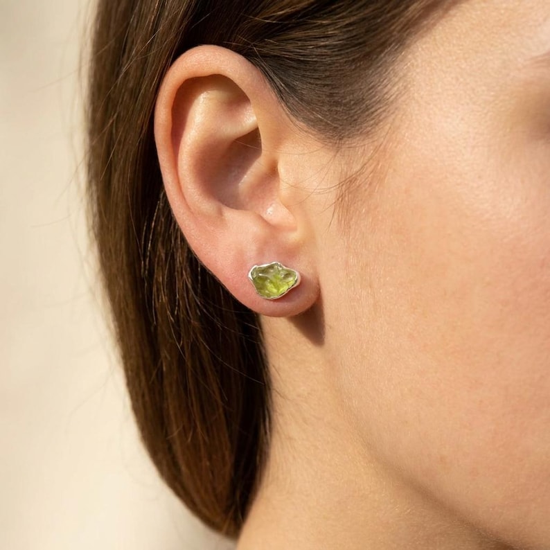 May include: A stud earring featuring a raw, irregular-shaped light green peridot gemstone set in a simple silver-toned metal bezel. The stone has a translucent, faceted appearance with natural inclusions and sits directly against the earlobe.