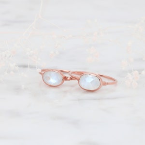 May include: Two rose gold rings featuring oval-shaped, faceted moonstones set in thin, polished metal bands. The stones exhibit a translucent, iridescent quality with subtle blue flashes, reflecting light across their multi-faceted surfaces.