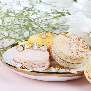 May include: A collection of gold rings featuring oval-shaped, iridescent moonstone cabochons. The rings are displayed on a white plate with a gold rim, resting atop pink, yellow, and beige macarons, with white flowers in the background.