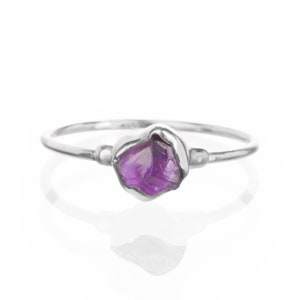May include: A silver ring featuring a raw, uncut amethyst gemstone. The purple gemstone is set in a bezel setting, with a simple band. The ring is displayed on a white background, highlighting the jewelry's natural beauty.