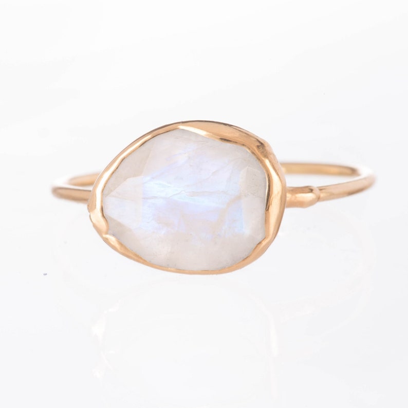 May include: A gold ring featuring a single, irregular, faceted moonstone set in a smooth gold bezel. The gemstone displays a translucent, milky white appearance with subtle blue iridescence. The band is thin, polished gold, creating a simple, refined look.