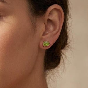 May include: A small stud earring featuring an irregular, organic-shaped lime green stone set in a polished gold-tone metal bezel. The earring is positioned on the earlobe, showcasing a smooth, rounded edge and a vibrant, translucent green hue.