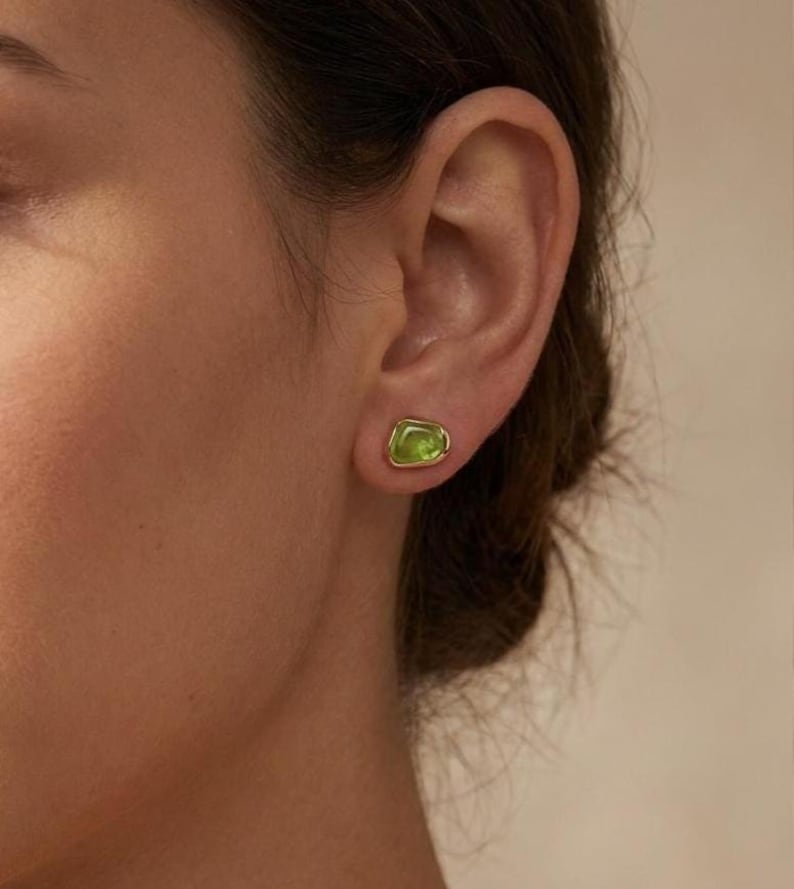 May include: A small stud earring featuring an irregular, organic-shaped lime green stone set in a polished gold-tone metal bezel. The earring is positioned on the earlobe, showcasing a smooth, rounded edge and a vibrant, translucent green hue.