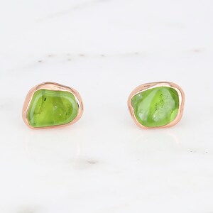 May include: A pair of stud earrings featuring irregular, polished lime green peridot gemstones set in smooth, rounded rose gold metal bezels. The stones have a translucent quality with natural internal inclusions visible within the green surface.