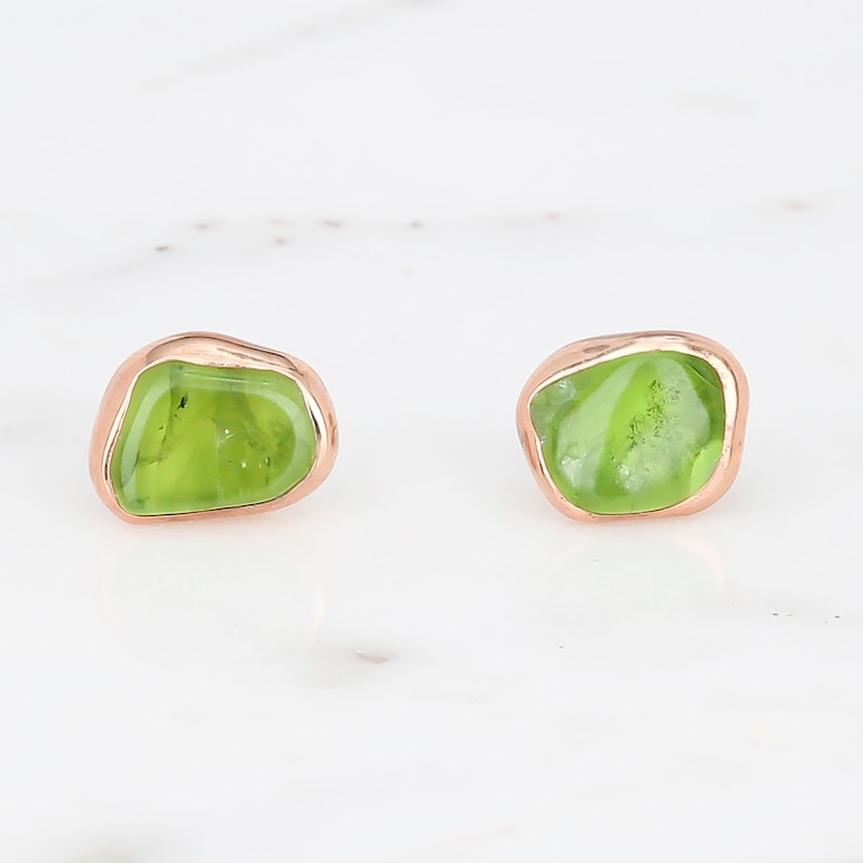May include: A pair of stud earrings featuring irregular, polished lime green peridot gemstones set in smooth, rounded rose gold metal bezels. The stones have a translucent quality with natural internal inclusions visible within the green surface.