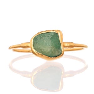 May include: A gold ring featuring a single, raw, uncut green gemstone set in a bezel. The band is thin and metallic gold, with a smooth, polished finish. The gemstone has an irregular, organic shape with a matte, textured surface in light green.