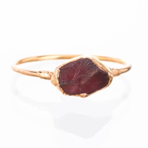May include: A gold ring featuring a raw, deep red garnet gemstone set in a textured metal bezel. The band is thin and polished, showcasing a natural, irregular shape for the stone with visible surface striations and a dark crimson hue.