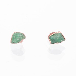 May include: A pair of stud earrings featuring raw, uncut green emerald stones. Each stone is encased in a metallic copper-colored setting that follows the irregular, jagged shape of the mineral. The earrings have a reflective, polished finish.