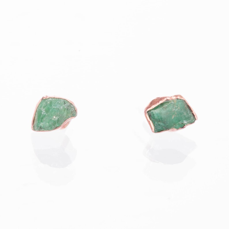 May include: A pair of stud earrings featuring raw, uncut green emerald stones. Each stone is encased in a metallic copper-colored setting that follows the irregular, jagged shape of the mineral. The earrings have a reflective, polished finish.