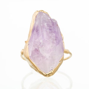 May include: A ring featuring a large, raw, light purple amethyst crystal set in a textured gold-toned metal bezel. The stone has a jagged, natural shape with visible facets and internal inclusions, mounted on a thin, smooth gold-toned metal band.